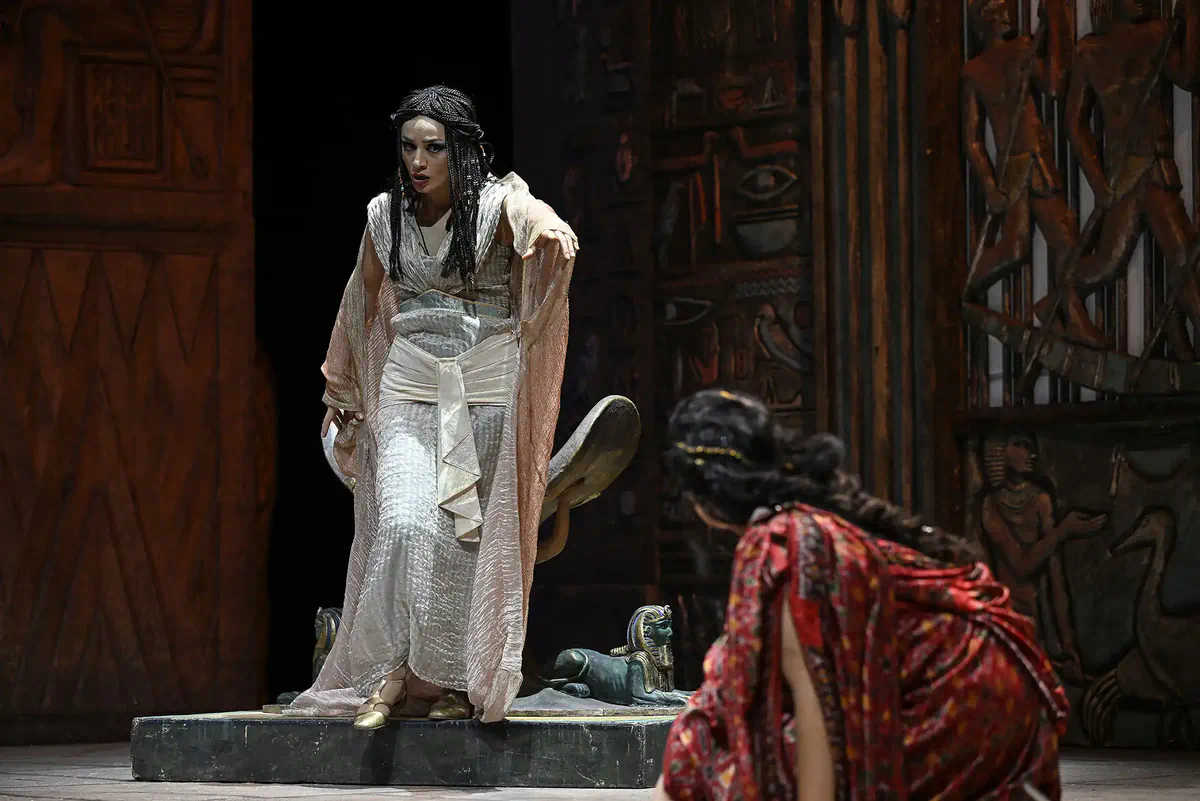 Escena de Aída con una mujer vestida de blanco sobre una plataforma y otra figura arrodillada en primer plano.