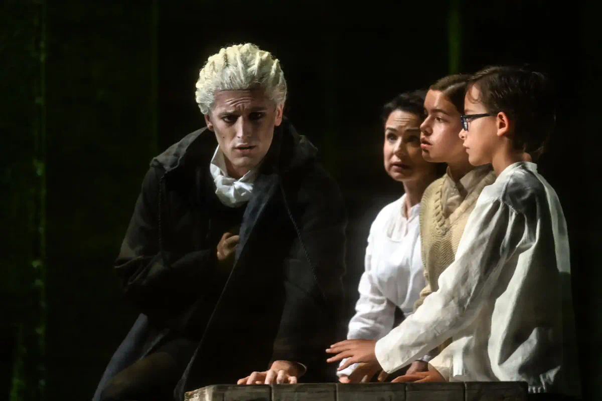Escena de La dama del alba con un intérprete de cabello blanco junto a tres personajes sentados.